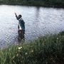 Ein Mann watet durch einen flachen, klaren Fluss, umgeben von grüner Vegetation am Ufer., CD