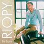 "RIOPY" und "Be Love" stehen links. Eine Person sitzt auf einem Klavier vor einem Fenster, in einem hellblauen Pullover., CD