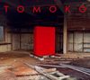 "TOMOKO" in großen roten Buchstaben. Im Hintergrund ein leerer, verlassener Raum mit einem roten Quader in der Mitte.