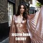 JOSEFIN ÖHRN CHERRY. Frau in schimmerndem, rosafarbenem Kleid, steht auf einer Straße zwischen Gebäuden., CD