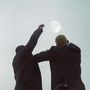 Zwei Personen in Anzügen stehen unter einem klaren Himmel und strecken die Arme zum Mond empor.