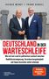 Titel: "DEUTSCHLAND IN DER WARTESCHLEIFE". Zwei Männer in Anzügen stehen vor Steinfiguren., Buch