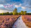 2026 WEGE PATHS CHEMINS. Ein Sandweg führt durch blühende Heide mit Bäumen im Hintergrund unter einem bewölkten Himmel.