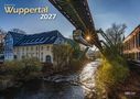 "Wuppertal 2027" steht groß im Bild. Eine Schwebebahn fährt über einen Fluss, daneben alte Gebäude und Bäume im Herbst.