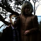 Eine Person trägt eine Schafmaske, neben ihr steht eine Frau mit blondem Haar und Mantel. Hintergrund: Bäume und Wasser.