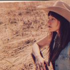 Ich kann keine Texte erkennen. Eine Frau mit Cowboyhut schaut nach links, vor einem trockenen Feldhintergrund.