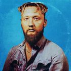 Ein Mann mit Bart und lockigem Haar trägt ein Jeanshemd vor einem blauen Hintergrund.