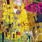 Graffiti mit dem Wort "kiss" in Pink über einem stilisierten Kuss-Paar in gelbmusterter Robe, umgeben von Blumen.