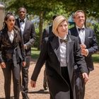 Ich kann keine Texte oder Namen im Bild erkennen. Eine Gruppe eleganter Menschen in formeller Kleidung läuft draußen.