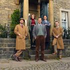 Fünf Personen in Vintage-Kleidung stehen vor einem Gebäude aus Stein mit Säulen und Sträuchern.