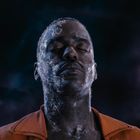 Ein Mann mit geschlossenen Augen, Gesicht teils mit frostähnlichem Muster bedeckt, vor einem dunklen, nebligen Hintergrund.