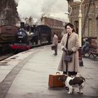 Aufschrift "RUSSELL" auf einem Schild. Eine Frau in einem beigen Mantel steht mit Koffer und Hund am Bahnhof. Eine Dampflok ist zu sehen.