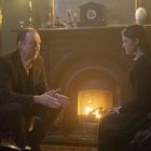 Ein älterer Mann und eine junge Frau sitzen in einem gemütlichen Zimmer vor einem prasselnden Kaminfeuer.