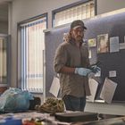 Ein Mann mit Baseballcap und Handschuhen steht an einer Pinnwand mit Fotos und Dokumenten in einem Büro.