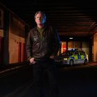 „Police“-Schrift auf einem Polizeiauto. Ein Mann in Lederjacke steht in einer dunklen, beleuchteten Unterführung.