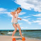 Ein Kind spielt am Strand, balanciert auf umgedrehten Eimern mit Seilen in den Händen, vor blauem Himmel und Wellen.