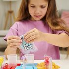 „Jelly Fun“ auf Tüte in Kinderhänden. Ein Mädchen in rosa Shirt spielt mit Bastelset am Tisch. Im Hintergrund ein Sofa.