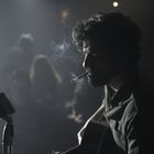 Ein Mann mit Bart spielt Gitarre, eine Zigarette im Mund. Ein altes Mikrofon ist in der Nähe, im Hintergrund unscharfe Personen.