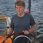 Ein älterer Mann steht am Steuer eines Segelbootes auf dem Meer, trägt ein graues T-Shirt. Im Hintergrund ist Wasser zu sehen.