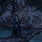 Eine Person in einem langen Mantel steht auf einem hölzernen Boot vor einer felsigen Küste bei Dämmerung.