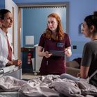 Eine Ärztin und zwei Frauen stehen um ein Krankenhausbett. Eine der Frauen hält ein Tablet, alle wirken nachdenklich.