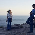 Keine sichtbaren Texte. Eine Frau und ein Mann, beide in Jeans, stehen an einer felsigen Küste mit Meer im Hintergrund.