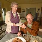 Ein älterer Raum mit Holzmöbeln. Eine ältere Frau reicht einem Mann eine Schüssel. Brot auf dem Tisch.