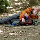 Ein Mann liegt in einem Wald auf dem Boden, trägt eine orangefarbene Jacke und hat eine Isomatte neben sich.
