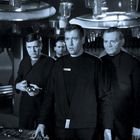 Vier Männer in dunkler Kleidung, ernst schauend, vor technischen Geräten in einem futuristischen Raum.