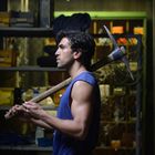 Keine Texte sichtbar. Ein Mann mit einem blauen Tanktop trägt eine Spitzhacke auf der Schulter in einer Werkstatt-Umgebung.