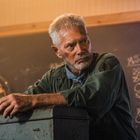 Ein Mann mit grauem Haar lehnt an einem Schrank. Im Hintergrund eine Dartscheibe und eine Tafel mit einer Liste.