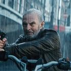 Keine sichtbaren Texte. Ein älterer Mann im Regen, hält eine Pistole, fährt ein Motorrad auf einer Brücke.