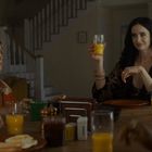 Eine Frau hebt ein Glas Orangensaft; um sie herum sitzen Menschen an einem Tisch mit Frühstück.