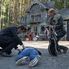 Ein Mann kniet neben einer am Boden liegenden Person. Im Hintergrund eine Band vor einem alten, hölzernen Haus im Wald.