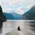 Kein Text erkennbar. Eine Person fährt auf einem Jetski durch einen malerischen Fjord, umgeben von hohen Bergen.