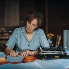 Eine Frau sitzt an einem Tisch und schreibt. Daneben steht eine alte Schreibmaschine. Im Hintergrund sind Bücher und Blumen.