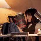 „Verity Crawford, Hospitality“ auf Buchcover. Frau sitzt entspannt mit Buch, Beine angezogen, Tisch voller Büroartikel.