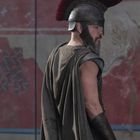 Kein Text vorhanden. Ein Mann in antiker Rüstung mit Helm und rotem Kamm, vor einem Wandgemälde stehend.