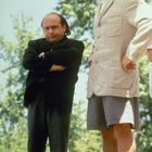Keine erkennbaren Texte. Zwei Männer stehen im Freien. Einer trägt einen Anzug, der andere eine helle Jacke und Shorts.