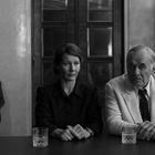 Eine Frau und ein Mann sitzen an einem Tisch mit Gläsern. Ein älterer Mann sitzt im Hintergrund. Schwarz-Weiß-Porträt.