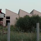 Ein Mann sitzt auf einem Dach, hinten graue Gebäude mit Sägezahn-Dächern. Eine Frau steht im Gras. Büsche und ein Zaun im Vordergrund.
