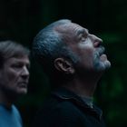 Zwei Männer im Wald, einer schaut nach oben. Der Hintergrund ist dunkel und bewaldet.