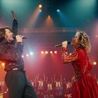 Zwei Menschen singen auf einer Bühne in schillernden Outfits. Bunte Lichter leuchten im Hintergrund, ein Chor applaudiert.