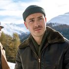 Zwei Männer in Winterlandschaft. Der Vordergrund zeigt einen mit Mütze und brauner Jacke, im Hintergrund schneebedeckte Berge.