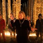 Fünf Personen stehen in einem Kreis aus Kerzen vor mystischen, bemalten Holzskulpturen in einem dunklen Wald.