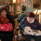 Zwei ältere Frauen auf Sesseln, eine öffnet eine rote Tasche, die andere ein silbernes Geschenkpapier. Wohnzimmerdekor.