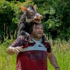 Eine verschmutzte Person trägt ein Wildschwein auf den Schultern in einer grünen, ländlichen Umgebung.