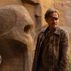 Ein Mann mit Augenklappe steht in einer felsigen Gegend. Im Hintergrund sind große Steinskulpturen in Form von Totenschädeln.