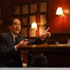 Eine Person sitzt an einer Bar, hält eine Zigarre und gestikuliert, umgeben von warmem Licht und gerahmten Bildern.