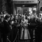 Der Text "PREMIERE" steht hinter einer elegant gekleideten Frau, umgeben von Fotografen und Reportern mit Blitzkameras.